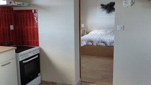 Een kleine keuken met een fornuis en een rode tegelwand, die uitkomt op een slaapkamer met een bed en nachtkastje.