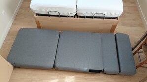 Een compact bed met houten frame en metalen hoofdbord, staand in een smalle kamer met houten vloer.