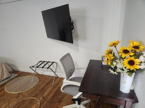 Una habitación con una televisión instalada en la pared, una mesita con un jarrón de girasoles, una silla y un taburete.