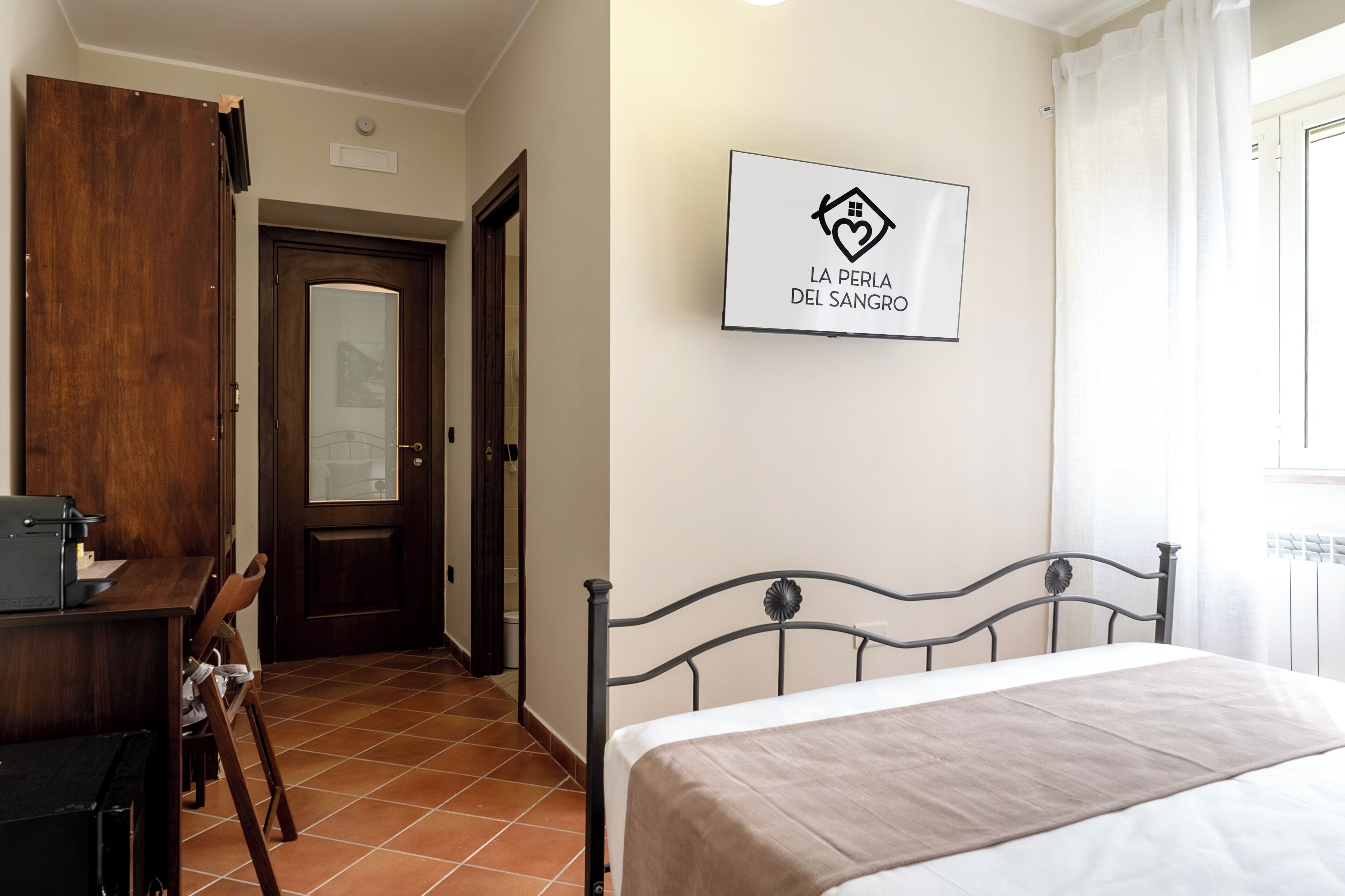 Una camera da letto con un letto, una scrivania e una sedia. Si vede una porta e una finestra con tende.