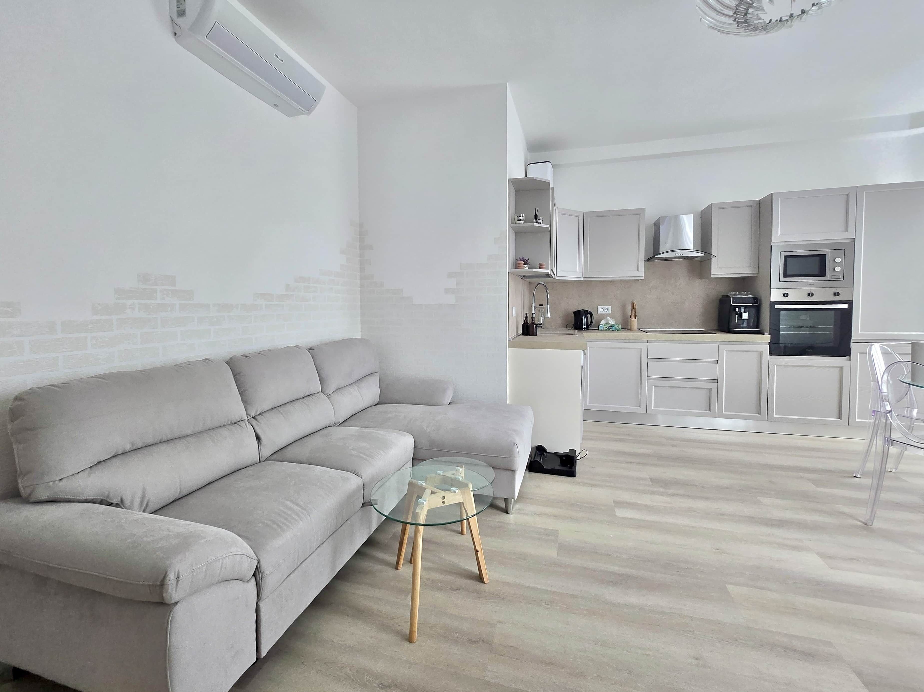 Un salon moderne avec un canapé gris, une table basse en verre et un coin cuisine comprenant des meubles blancs, un four à micro-ondes et un four traditionnel.