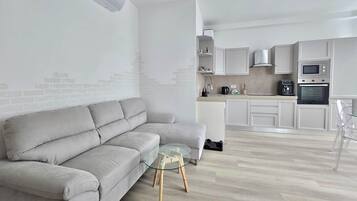 Un salon moderne avec un canapé gris, une table basse en verre et un coin cuisine comprenant des meubles blancs, un four à micro-ondes et un four traditionnel.