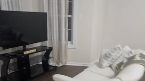 Una sala de estar con un sofá blanco, un televisor de pantalla plana sobre un mueble, una ventana con cortinas y suelo de madera.