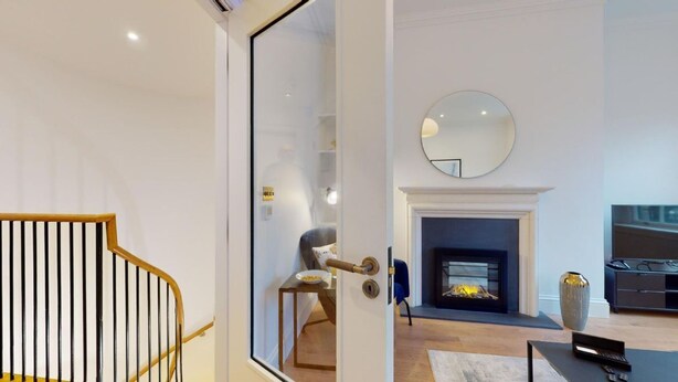Een moderne woonkamer met een open haard, een ronde spiegel aan de muur en een trap met een glazen deur naar een andere kamer.