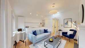 Een moderne woonkamer met een bank, een salontafel en een blauwe fauteuil. Achterin bevindt zich een keuken met een wit aanrecht en een spoelbak.