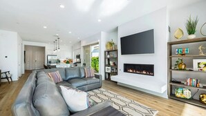 A modern living room with a grey sectional sofa, a fireplace, a flat-screen TV, and a bookshelf. - Platinum Luxury @ PGA West (La Quinta)