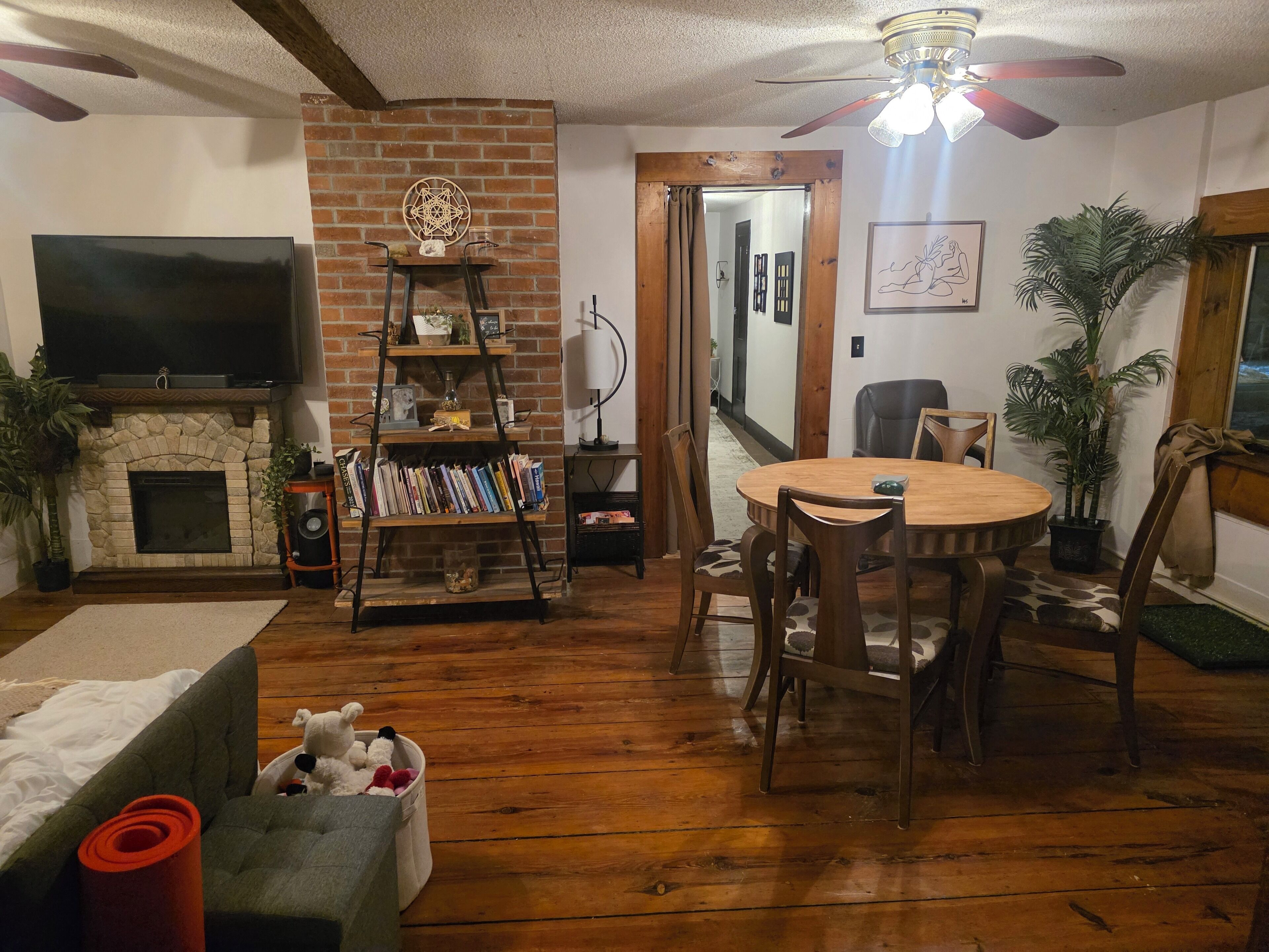 A living room with a fireplace, a TV mounted on the wall, a wooden dining table with chairs, a bookshelf, and a ceiling fan.