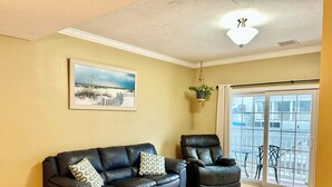 Ein Wohnzimmer mit schwarzem Ledersofa, einem schwarzen Ledersessel, einem kleinen runden Tisch, einer hängenden Pflanze und einem großen Fenster mit Blick auf einen Balkon.