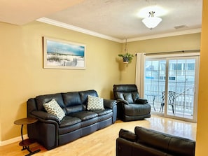 A living room with a black leather sofa, a black leather armchair, a small round table, a hanging plant, and a large window with a view of a balcony. - Spacious 2-bedroom, 2 bathroom, 2 car parking just steps to beach. (Hampton)
