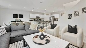 Una sala de estar moderna con un sofá seccional gris, una mesa de centro blanca y una cocina al fondo.