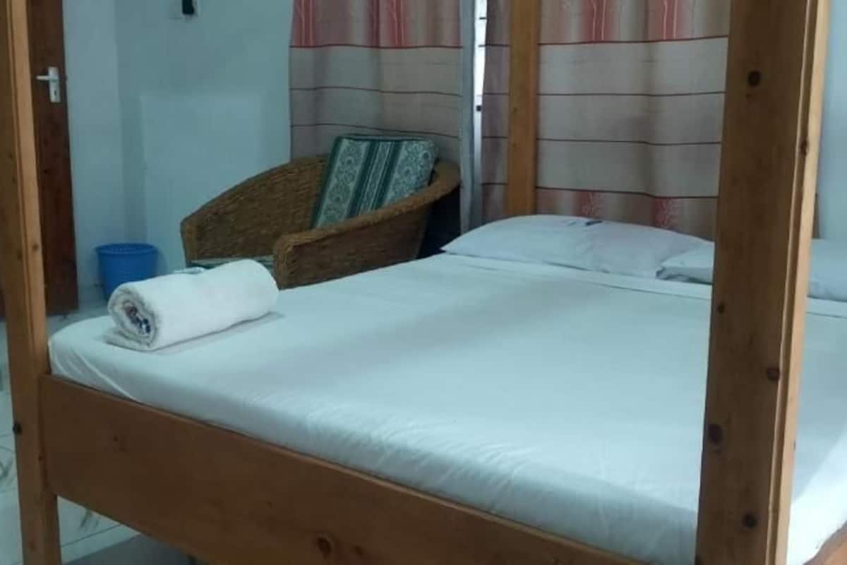 A single bed with a wooden frame, a blue trash bin, and a wicker chair with a cushion.