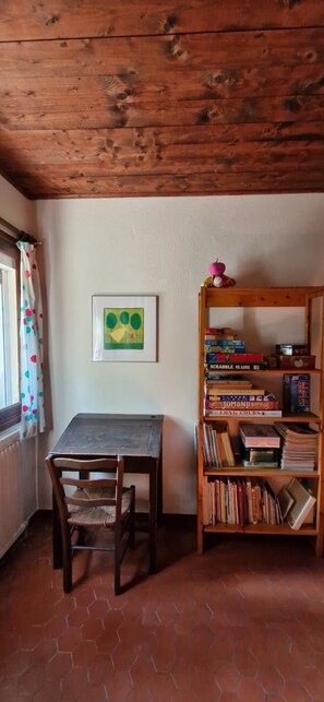 A room with a wooden table and chair, a bookshelf with books and games, a framed picture on the wall, and a window with a curtain. - Beautiful family home with sea view (Carqueiranne)