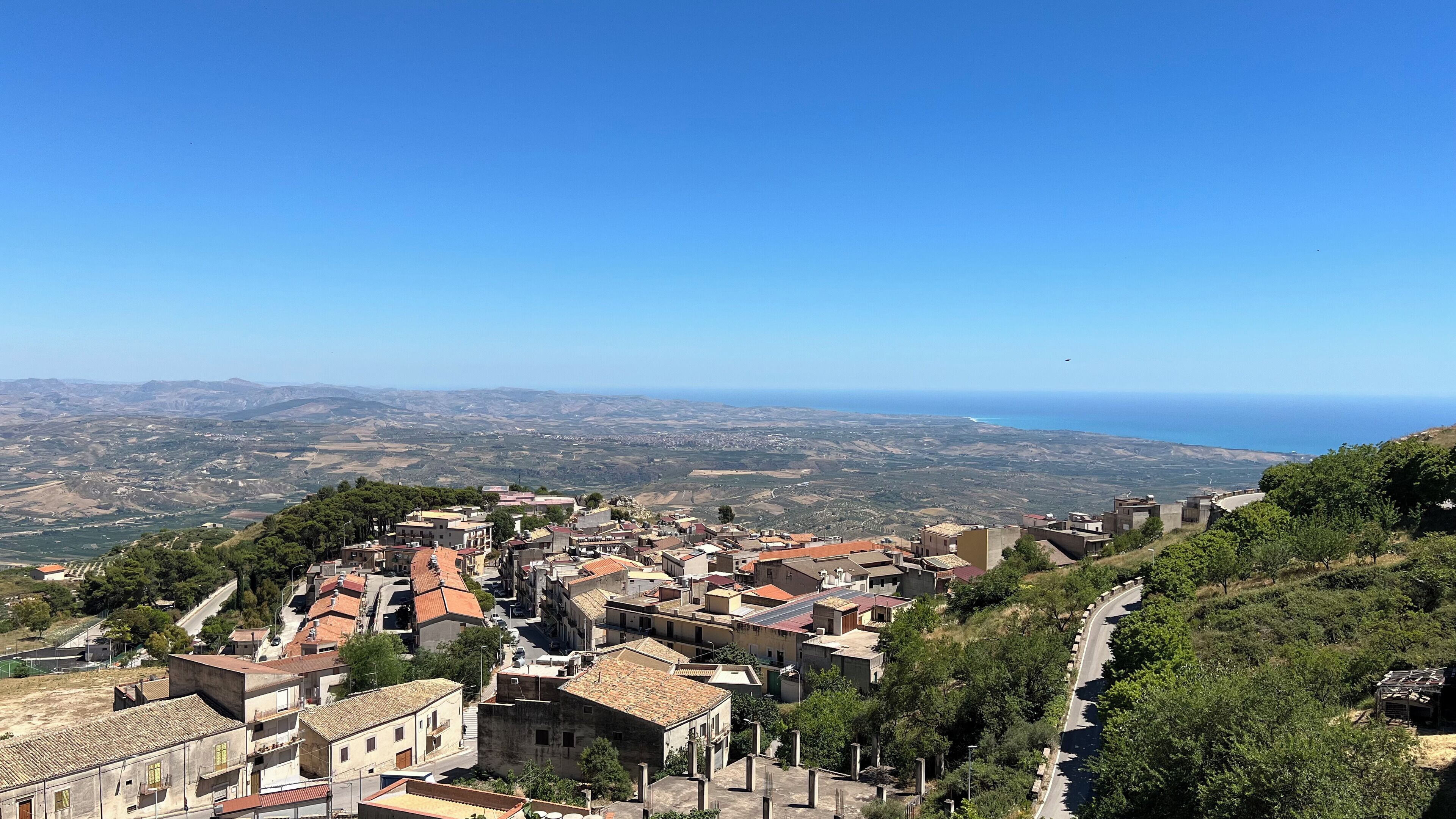 A coastal town with terracotta-roofed buildings, a winding road, and a view of the sea and distant hills.