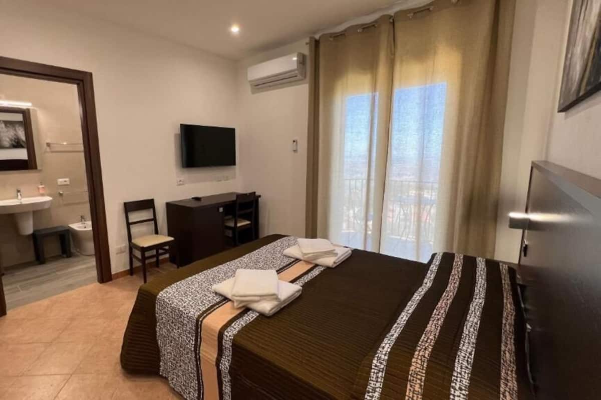A hotel room with a bed, a desk, a chair, a TV, a bathroom with a sink, and a window with curtains.