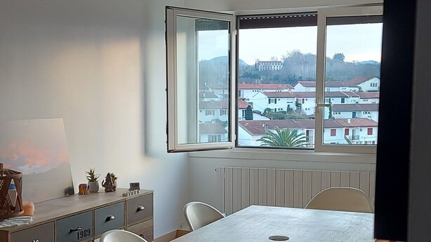 Ein geräumiger Raum mit großem Esstisch, weißen Stühlen, einem Holzschrank und Blick auf Häuser durch ein großes Fenster.