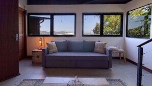 A modern living room with a grey sofa, a small side table with a lamp, and large windows offering a view of the outdoors. - Loft by the Lagoon: Bathtub with View & Nature (Florianópolis)