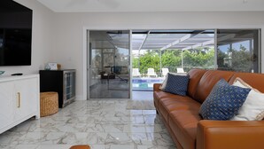 A modern living room with a leather sofa, a flat-screen TV, and a glass door leading to a pool area. - Waterfront Canal Home with Private Pool . Family Getaway . New Rental (Cape Coral)
