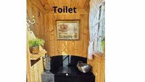 L’intérieur d’une cabane en bois comprend des toilettes, un tableau encadré accroché au mur, une étagère avec une plante et un banc noir sur lequel se trouve un panier.