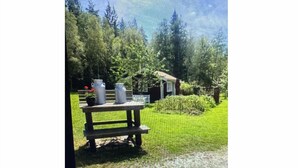 Une table de pique-nique en bois, avec des récipients en métal, et une petite maison dans un espace verdoyant bordé d’arbres.