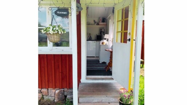 Une véranda en bois avec une pancarte d’accueil, des fondations en pierre et une vue sur l’espace cuisine.