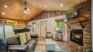 A cozy living room with a fireplace, leather sofa, and a television mounted on a stone wall. - Time Falls Cabin | 2 King BR | 2.5 BA | 2 Porches | Fire Pit | Fast WiFi (Mentone)