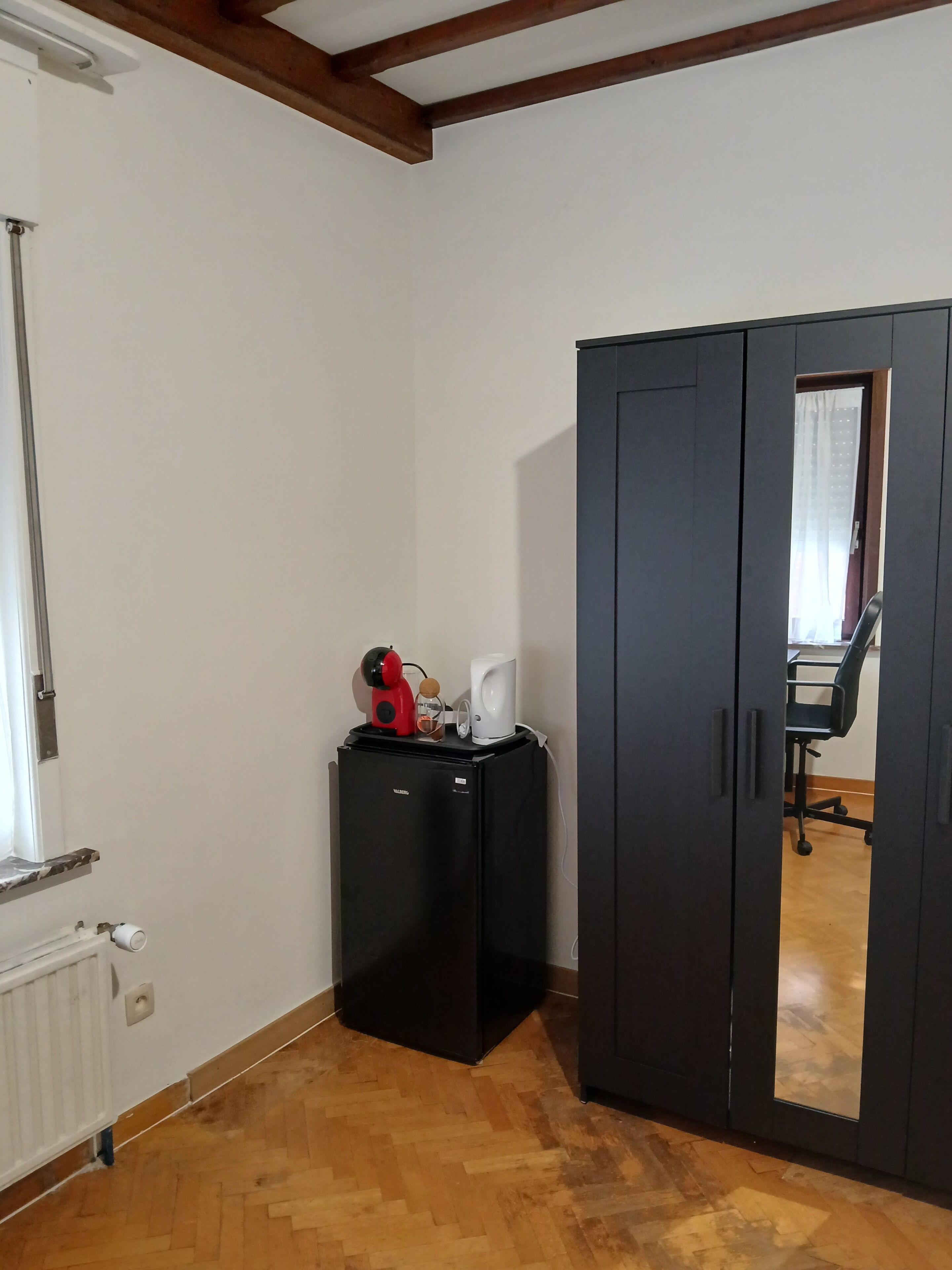 A room with a black wardrobe, a small black refrigerator, a radiator, and a wooden floor.