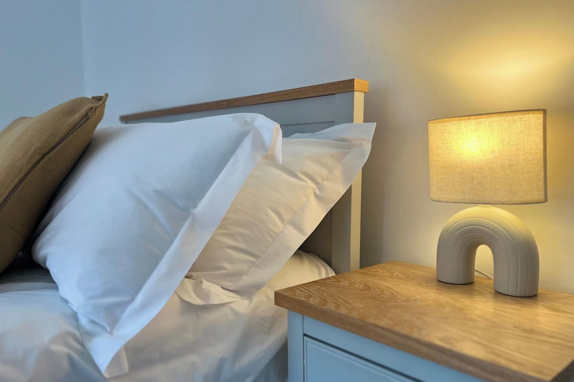 Una cama bien hecha con ropa blanca, un cabecero de madera y una mesita de noche con una lámpara.
