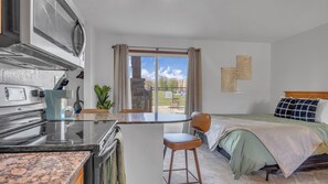 Une chambre d’hôtel compacte dotée d’une kitchenette, d’un lit recouvert d’une courtepointe verte, d’une tête de lit en bois et donnant sur un espace extérieur clos.