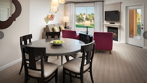 Ein Hotelzimmer mit Esstisch, Stühlen, Obstschale, einer Couch, einem Fernseher, einem Kamin und einem großen Fenster mit Blick auf Palmen.