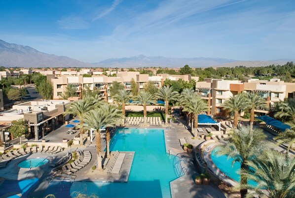 Een resort met een centraal gelegen zwembad, palmbomen en een bergachtige achtergrond.