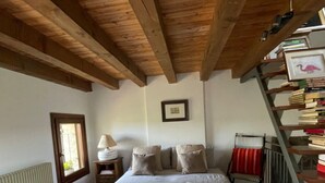 Une chambre avec un plafond en bois, un lit recouvert de draps blancs, une table de chevet avec une lampe, un radiateur, une fenêtre et une bibliothèque.