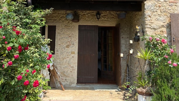 Un bâtiment rustique en pierre, doté d’une porte et de volets en bois, entouré d’une végétation luxuriante et agrémenté de plantes en pot.