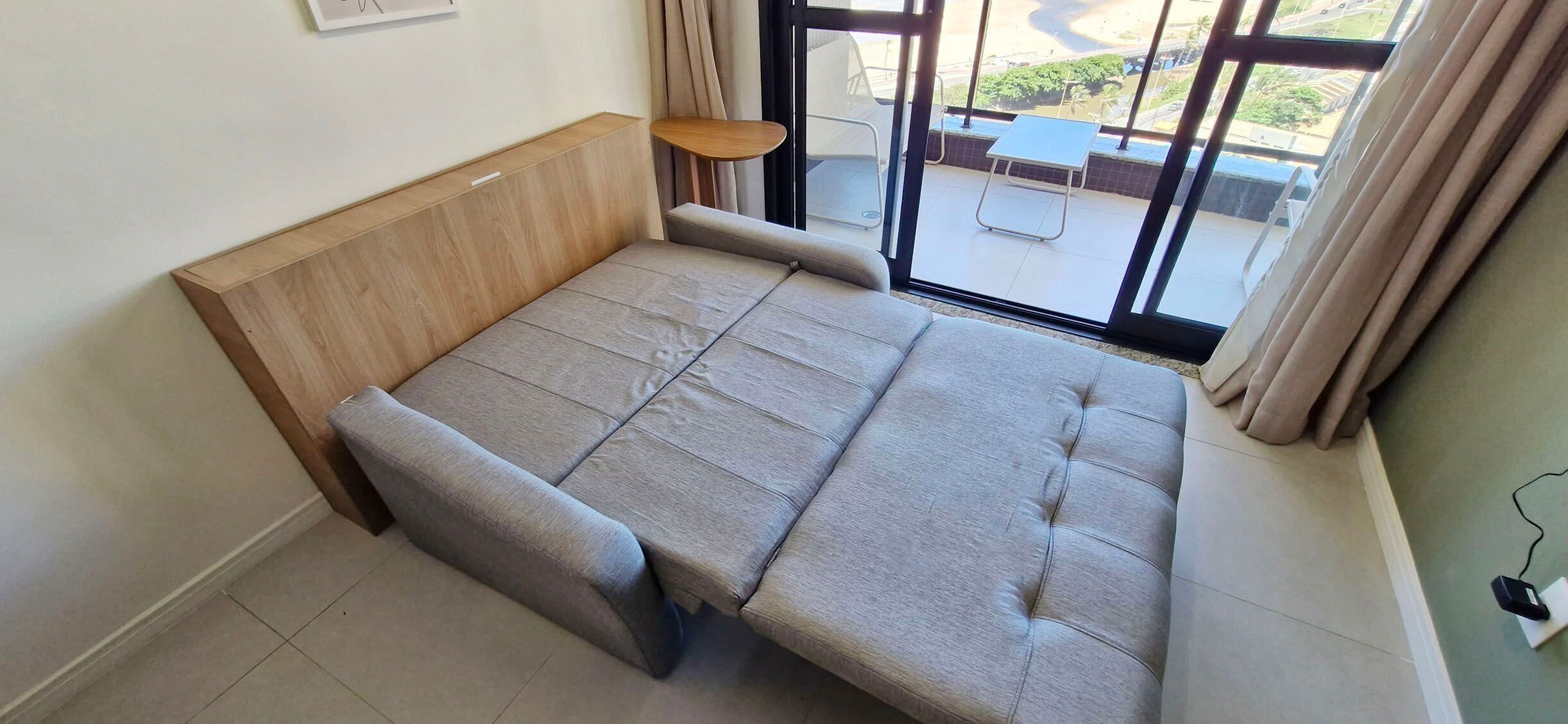 A room with a sofa bed, a small side table, and a view of the outside through a large window.