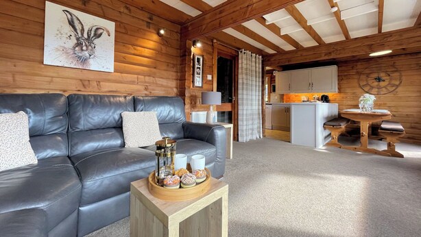 L’intérieur chaleureux d’un chalet en rondins, avec un canapé en cuir, une table basse en bois sur laquelle sont disposés des en-cas, un coin cuisine et une horloge au mur.