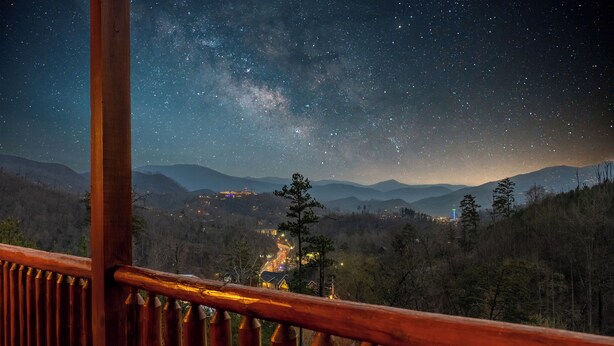 Nachtsicht von einem Holzbalkon mit Blick über ein Tal, einen sternenklaren Himmel und ferne Berge.