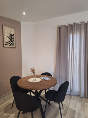 Un coin repas avec une table ronde en bois, quatre chaises noires, un vase contenant des plantes séchées et une œuvre d’art encadrée accrochée au mur.