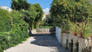 Un sendero de grava conduce a un edificio con balcón, rodeado de abundante vegetación y una valla.
