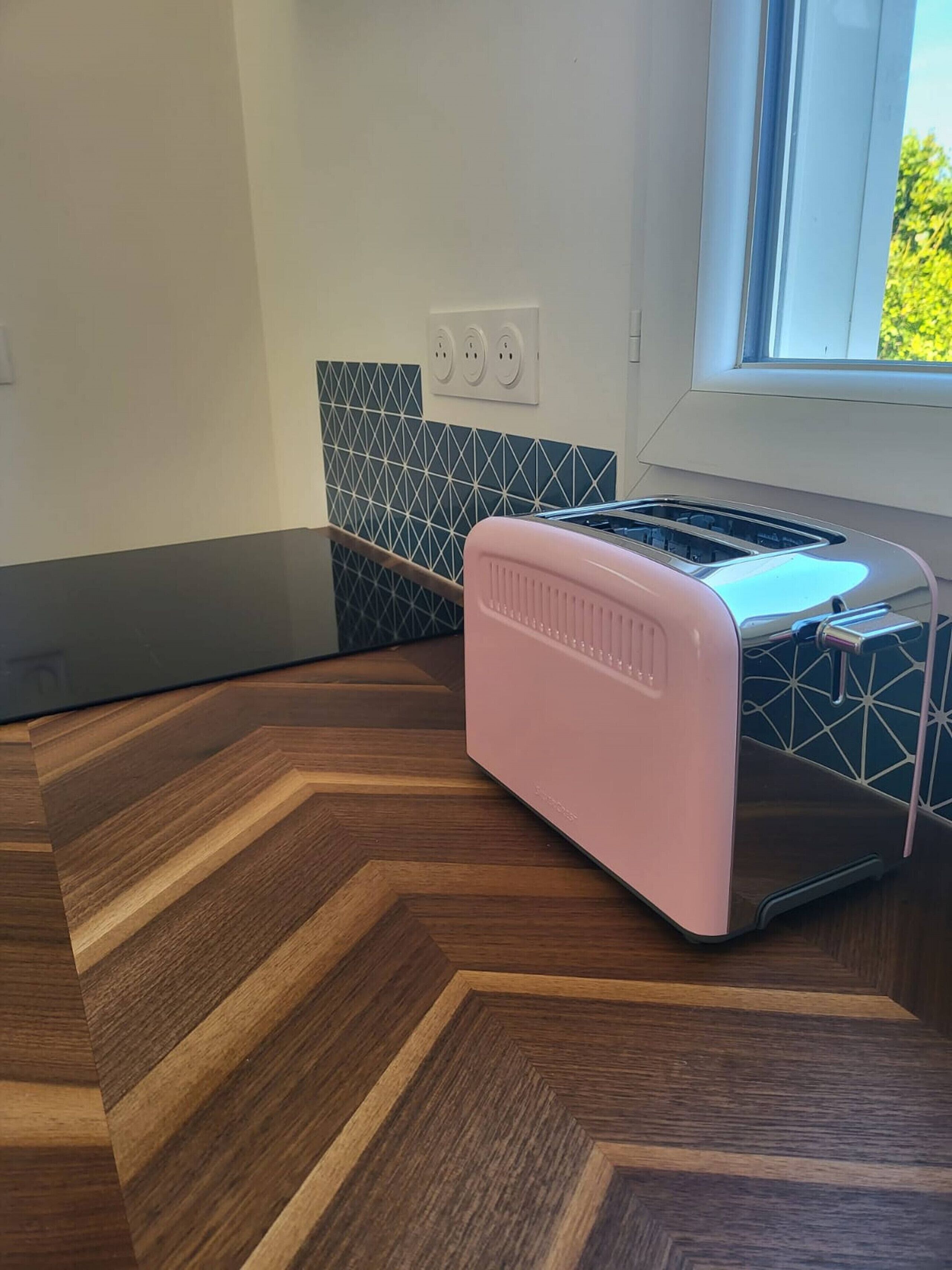 Una cocina con un tostador rosa sobre un piso de madera con patrón de espiga, un salpicadero de azulejos blanco y negro, y una ventana con vistas a la vegetación.