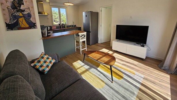 Una sala de estar moderna con un sofá, una mesa de centro y un televisor. Hay una zona de cocina con un refrigerador, un lavaplatos y una ventana.
