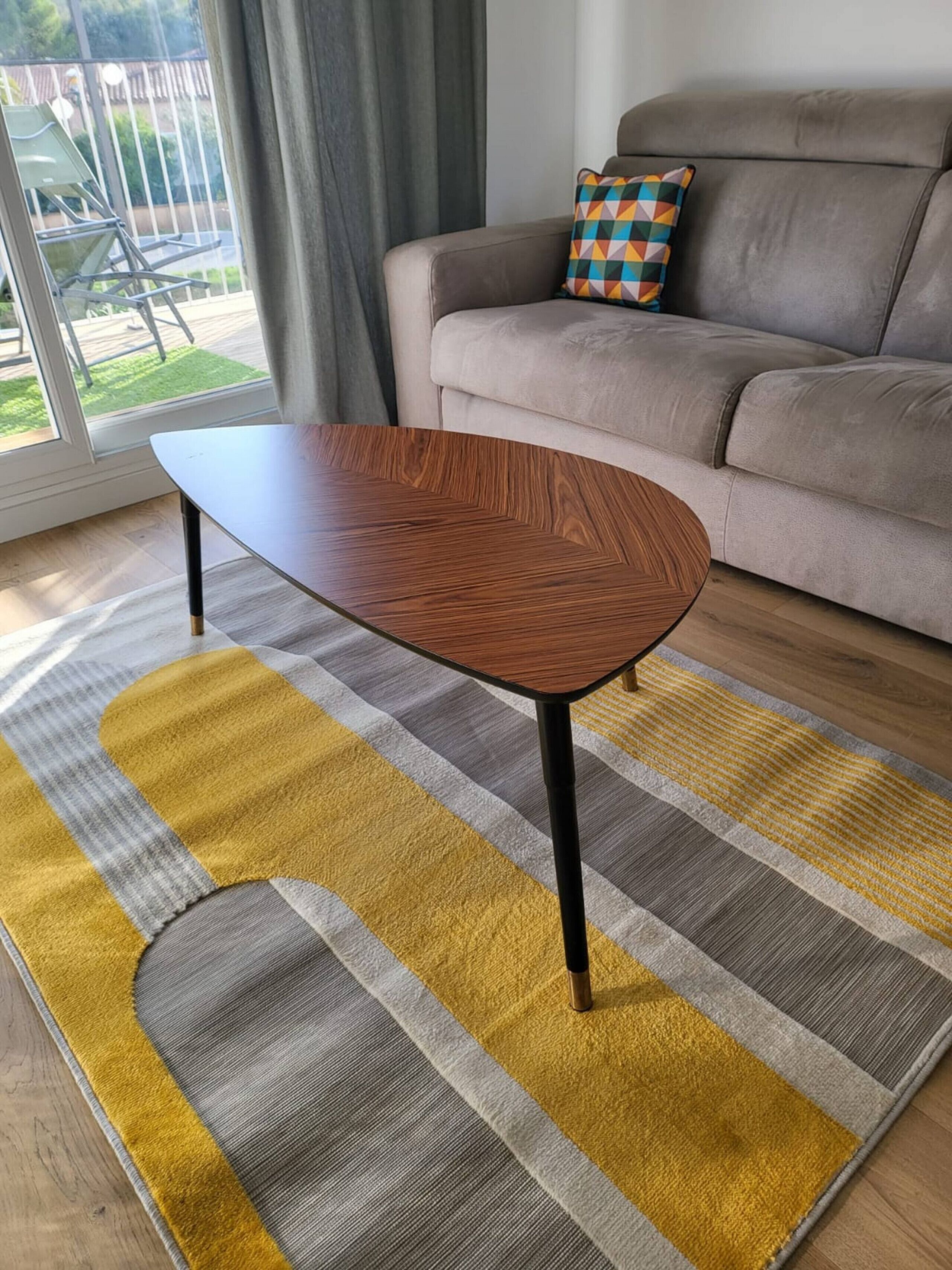 Un salon moderne avec une table basse en bois, un tapis à motifs rayés et un canapé gris agrémenté d’un coussin coloré.