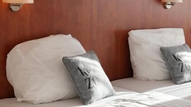 A neatly made bed with white linens and two pillows, one with a 'Z' design, in a room with wooden headboard and wall-mounted lamps. - Gästehaus Tilly Hotel (Hann. Muenden)