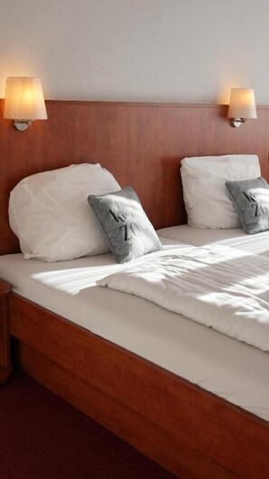 Una cama bien tendida con sábanas blancas y dos almohadas, una con diseño en forma de 'Z', en una habitación con cabecera de madera y lámparas de pared.