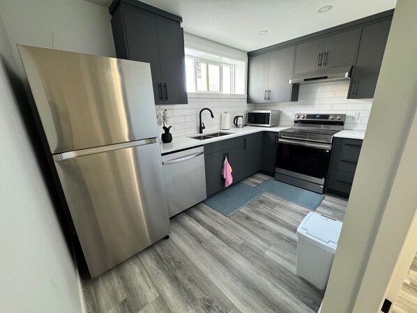 A modern kitchen with stainless steel appliances, dark cabinets, and a window providing natural light. - The Secure Haven (Regina)