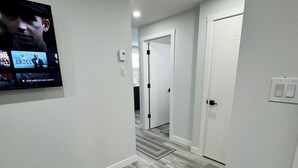 A modern hallway with a flat-screen TV displaying a show, light-colored walls, and a grey carpet. - The Secure Haven (Regina)