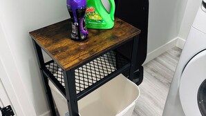A laundry station with a washing machine, a green detergent bottle, and a purple spray bottle on a wooden table. - The Secure Haven (Regina)