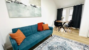 Un soggiorno moderno con un divano blu, due cuscini arancioni, un tappeto decorato e un'area pranzo con un tavolo e sedie neri.