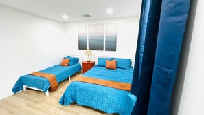 Una habitación con dos camas, cada una con ropa de cama azul y almohadas decorativas naranjas, una mesita de noche con una lámpara, y un ventanal con vista al mar.