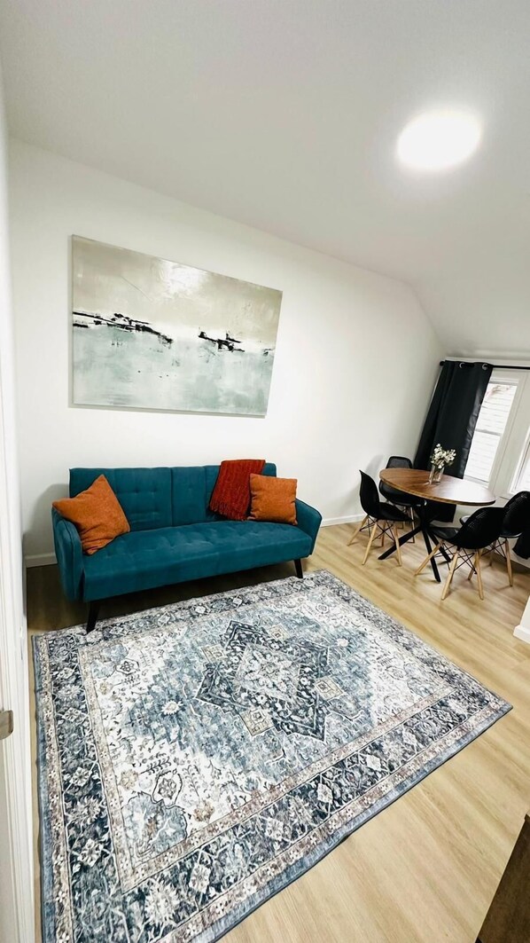 Una sala de estar con un sofá azul turquesa, un tapete estampado, un cuadro en la pared y una zona de comedor con una mesa y sillas.