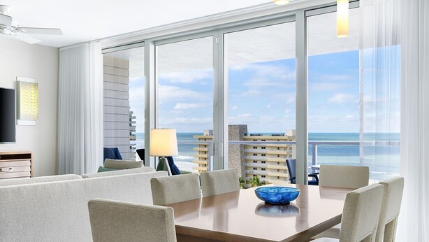 A modern hotel room with a large balcony, a dining area with a table and chairs, and a television on a stand.