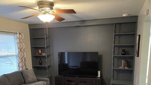 A living room with a dark wood floor, grey sofas, a flat-screen TV, and built-in shelves.